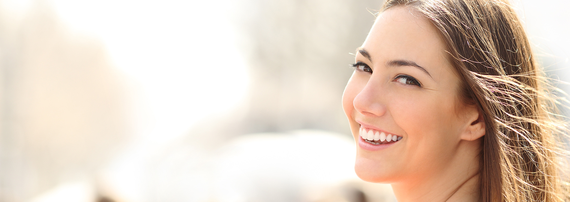 The image shows a woman smiling at the camera with her hair blowing in the wind, set against a blurred background.