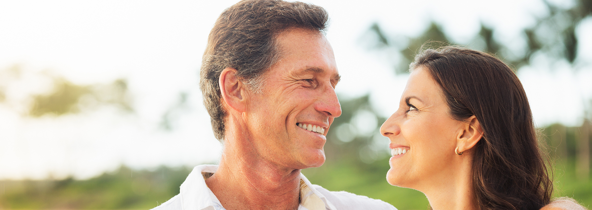 The image shows a man and woman smiling at each other with their faces close together they appear to be standing outdoors during daylight, possibly on a beach.
