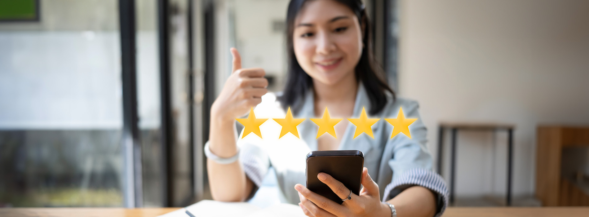 The image shows a person taking a selfie with a smartphone while holding up a thumbs-up gesture. The individual appears to be indoors, possibly at a cafe or similar setting, with a blurred background that suggests a casual atmosphere.