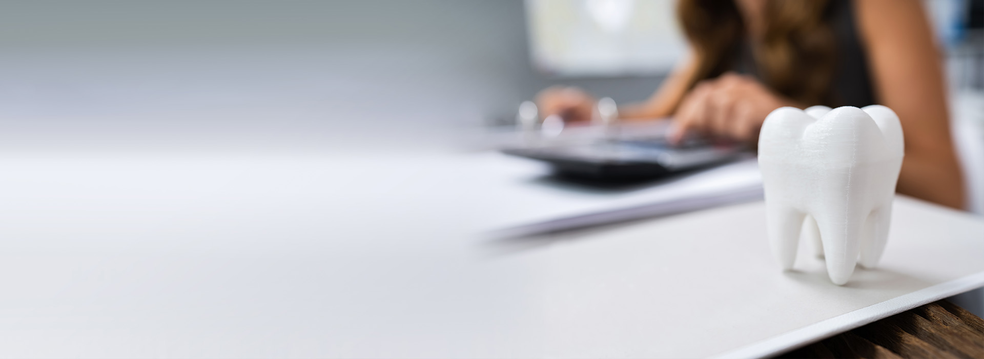 The image shows a person sitting at a desk with a computer monitor, surrounded by office items, while a small white toothbrush-shaped object is prominently placed on the desk.