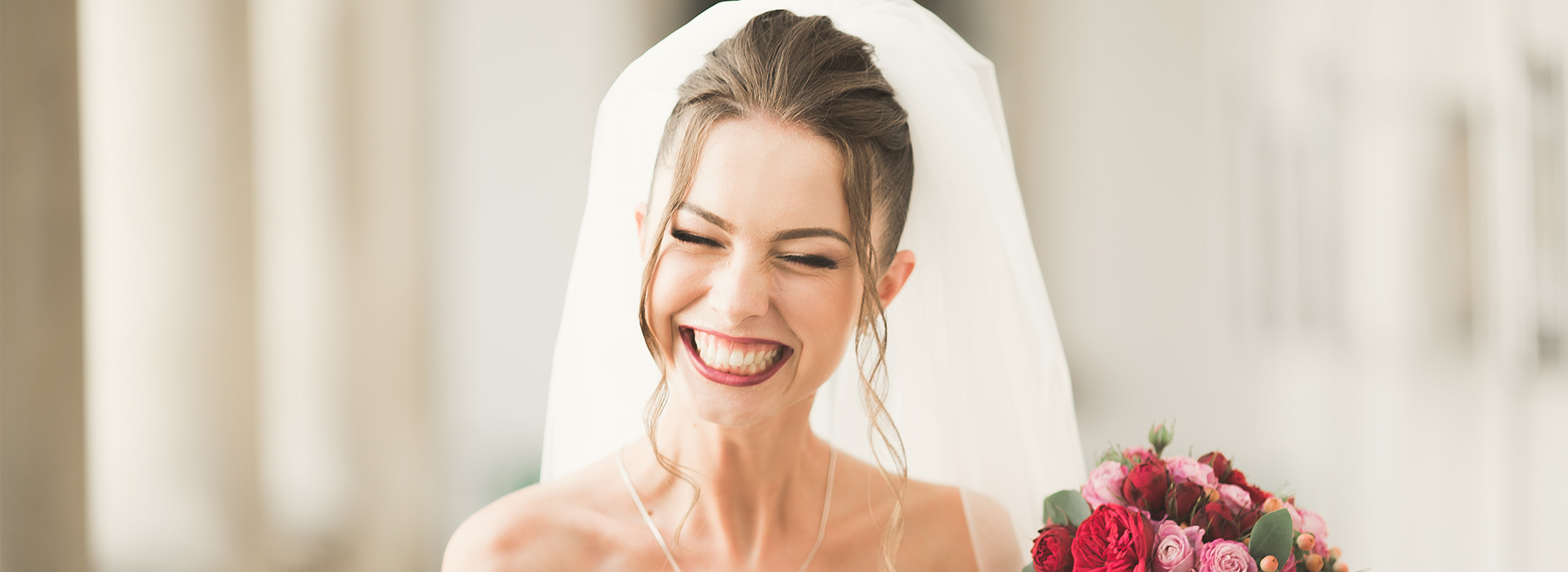 The image shows a woman wearing a white wedding veil and a bridal necklace, standing next to a bouquet of flowers, with a smile on her face.