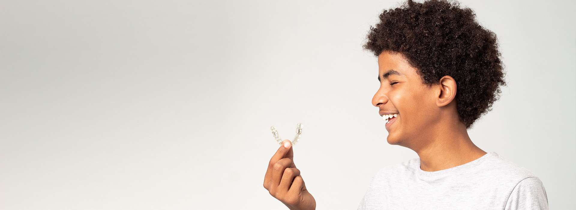 The image features a young person with curly hair holding a small object in their hand, smiling at the camera against a plain background.