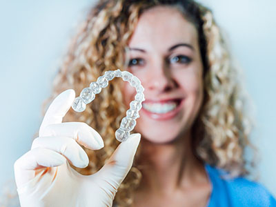 The image features a smiling woman holding up a clear dental retainer with her left hand.