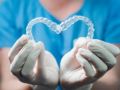 This image features a human hand holding a heart-shaped object made of clear plastic material, which appears to be a dental appliance such as a retainer or mouthguard, against a background that suggests a medical setting with blue and white colors.