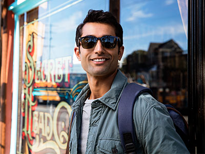 The image features a man standing outdoors, wearing sunglasses, a jacket, and holding a backpack, posing for a photograph with a smile against a backdrop of a storefront window.