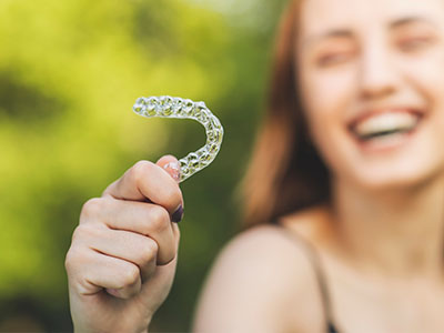 A smiling woman holding up a clear plastic retainer with her left hand.