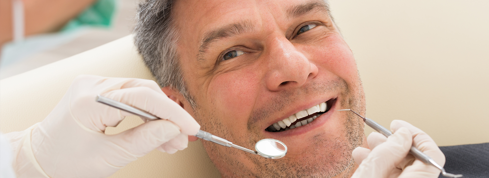 A man with a toothy grin receiving dental care from a professional, with a visible dental chair and tools in the background.