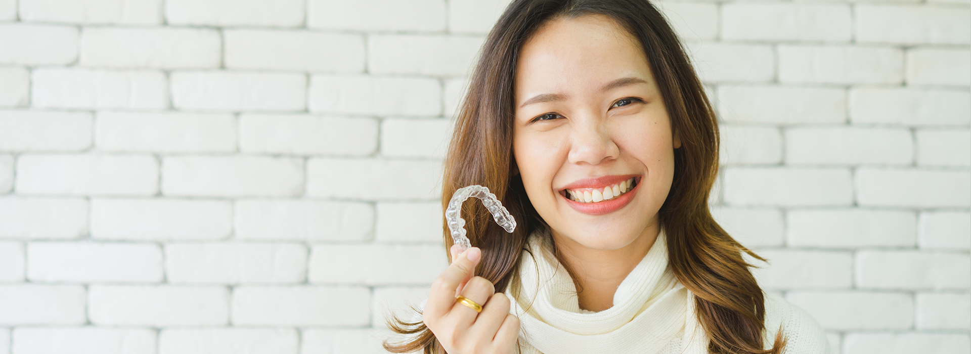 A smiling woman holding a toothbrush with her mouth while standing in front of a brick wall.