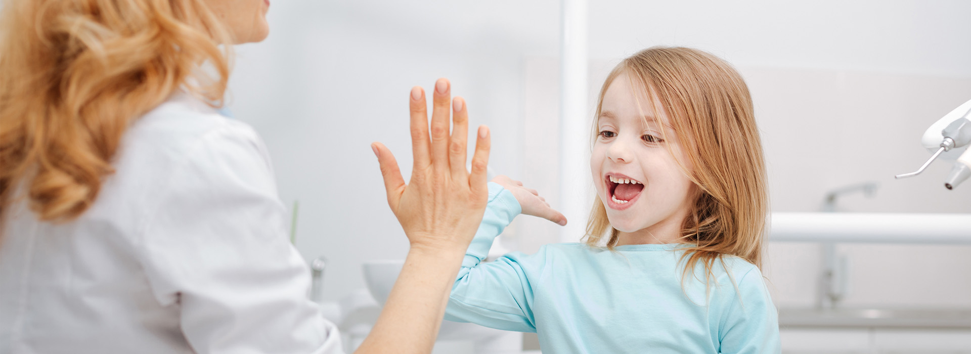 A young girl with blonde hair is waving at her mother who is washing dishes in the background.