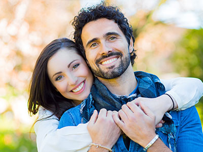 A man and woman embrace each other outdoors during daylight, with the man sporting a beard and both smiling.