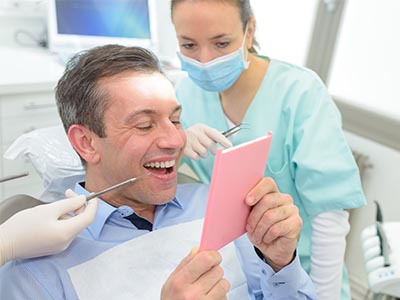 The image shows a man sitting in a dental chair with a smile, holding up a pink cardboard sign with his mouth, while a female dental professional stands behind him, observing.