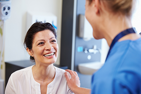 The image shows two individuals in a medical setting on the left, there s a woman wearing a white shirt and a necklace, smiling at another person, who appears to be a healthcare professional in scrubs, with both of them engaged in a conversation.