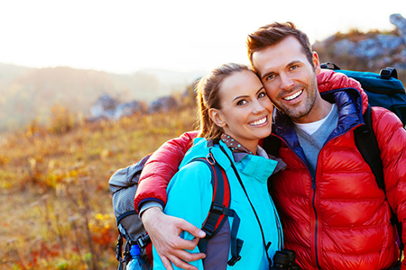 A man and woman smiling at the camera while standing outdoors during daylight, with the man wearing a backpack and both dressed for hiking or outdoor activities.