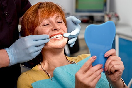 The image shows a woman sitting in a dental chair with a blue mouthguard on her teeth, holding up a mirror to show her teeth being worked on by a dentist who is smiling at the camera.