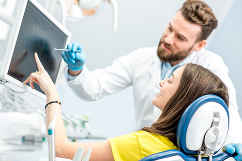 The image shows a man and woman in a dental office setting, with the man standing by a patient who is seated in a dental chair, both looking at a computer screen with the man pointing at something on it.