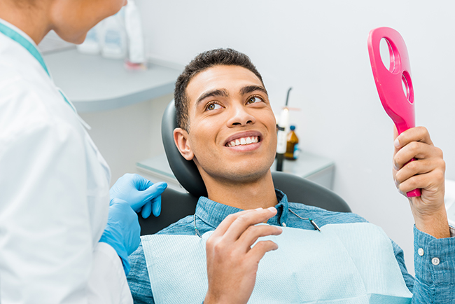 A young man is sitting in a dental chair with a smile on his face while receiving dental care from a professional wearing a blue uniform and holding a pink object.