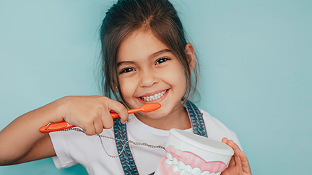 A young girl brushing her teeth with an oversized toothbrush while holding a toothpaste tube.
