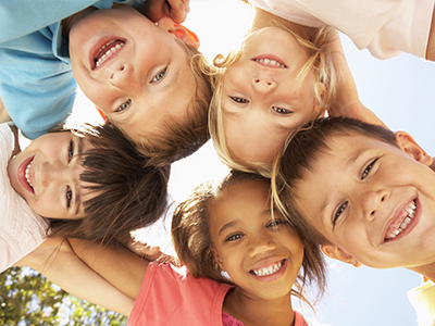 The image shows a group of children posing together with smiles on their faces, all looking towards the camera, against a clear sky background.