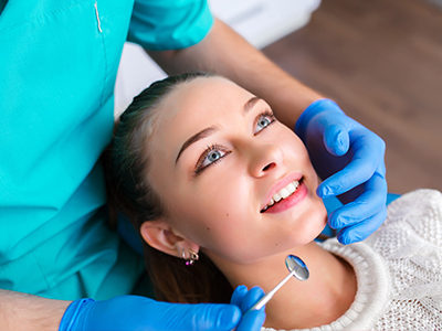 In the image, a woman is sitting in a dental chair with her eyes closed, receiving dental care from a professional who is using a dental tool on her teeth.
