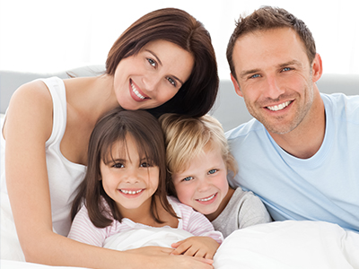 The image depicts a family of four people posing together on a bed with smiles and casual attire, with a man and woman facing forward and two children lying down behind them.