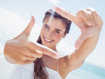 The image shows a woman taking a selfie with her hand held up against the camera lens, smiling at the camera. She has long hair, is wearing a white top, and appears to be outdoors during daylight.